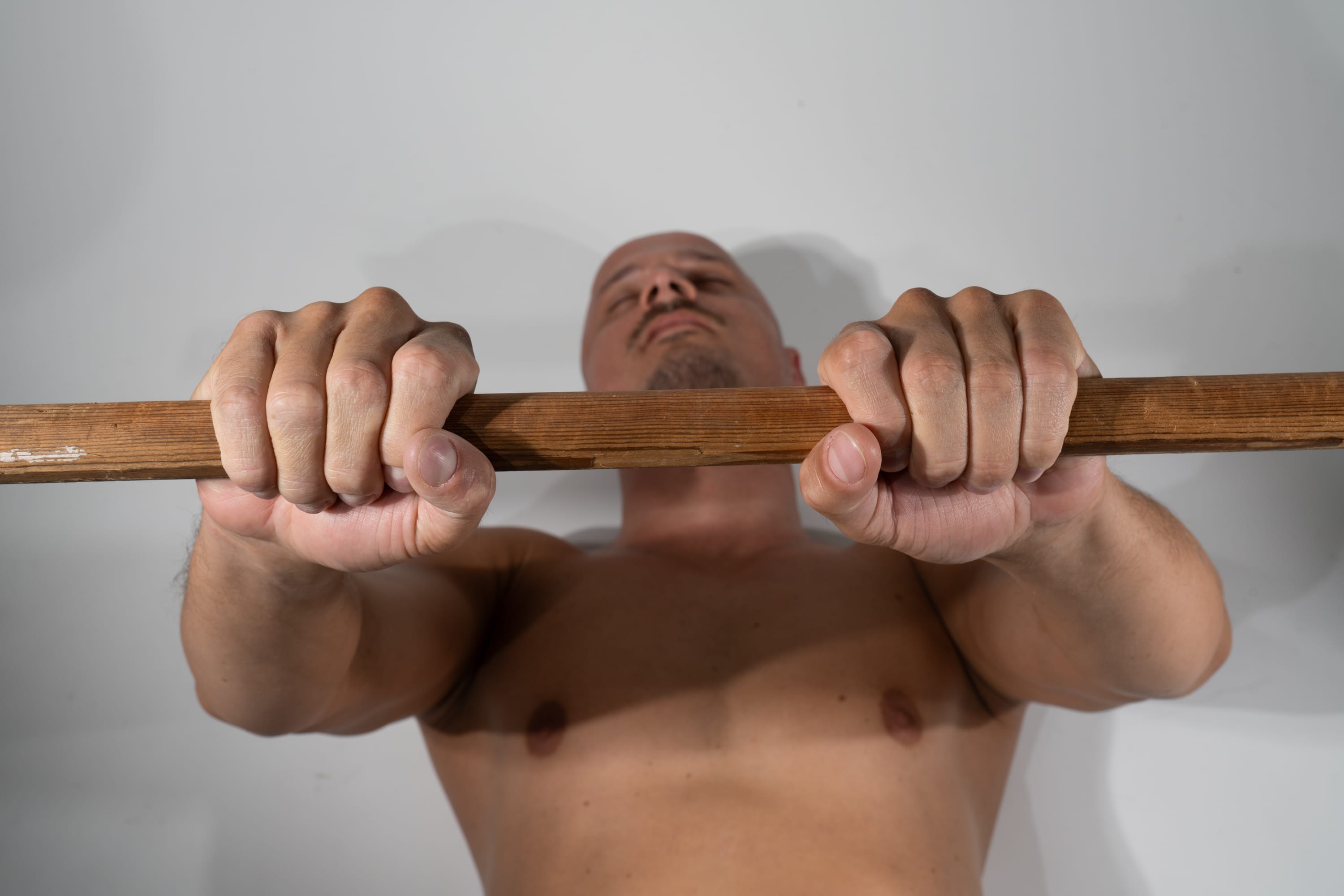 a shirtless, muscular man is shown from a low angle perspective, gripping a wooden bar as if about to perform a pullup. he is set against a plain white background. his hands are clasped firmly around the bar, his face turned upwards with a focused expression. the lighting is soft and natural, highlighting the definition of his muscles.
