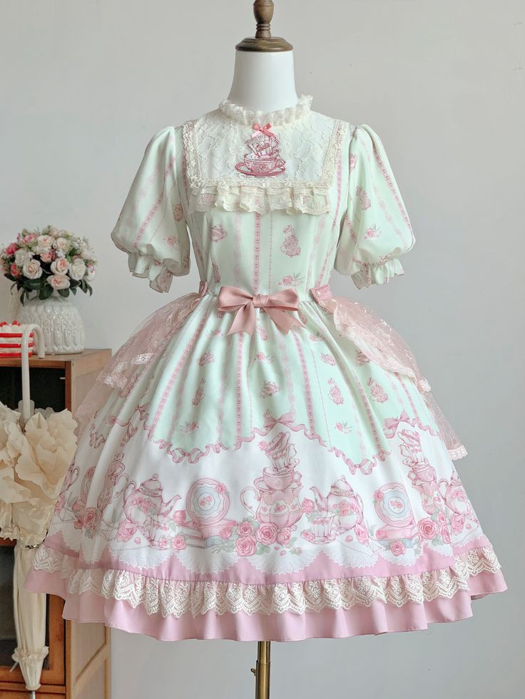 a light green and pink lolita dress is displayed on a mannequin. the dress features a print of cakes and flowers, with lace and ribbon details. the dress is shown in front of a white wall, with a vase of flowers and a wooden cabinet in the background.