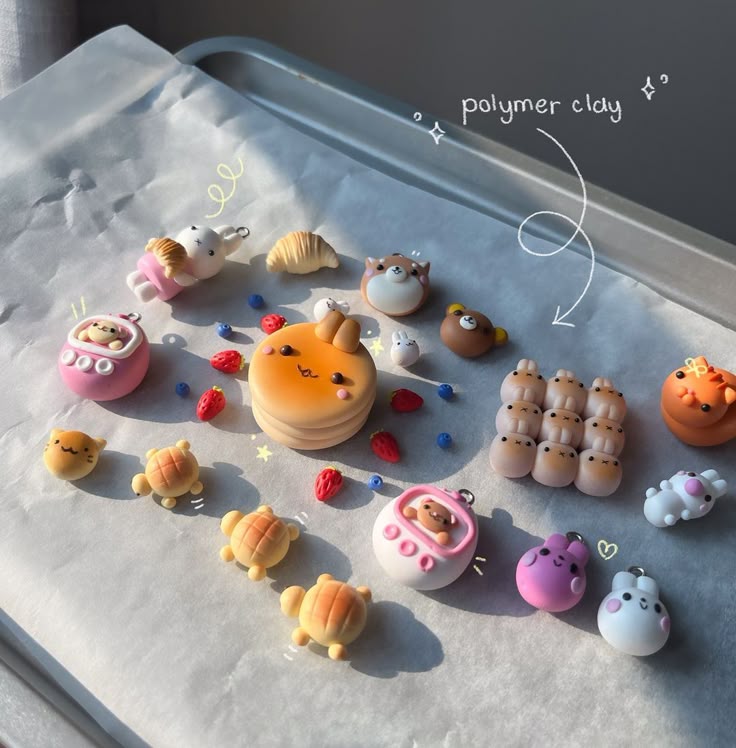 the image displays a collection of handmade polymer clay charms arranged on a baking tray lined with parchment paper. the charms are shaped like various objects, including food items like pancakes, a croissant, strawberries, and blueberries, as well as adorable animals such as bears, rabbits, turtles, and cats. there are also charms that resemble tamagotchi toys. the items are rendered in a kawaii style with pastel colors, and an arrow points to the collection, labeled as "polymer clay".