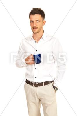 a man in a white shirt and beige pants is standing in front of a white background, smiling and holding a blue coffee mug. he is casually dressed and looks confident.