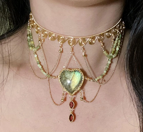 a closeup selfie showcasing a woman wearing an intricate gold choker necklace. the necklace features a prominent heartshaped green gemstone in the center, from which delicate chains with small red gemstones dangle. the choker band itself is adorned with spiraled details and connected to strands of smaller green gemstones on either side. the overall aesthetic leans towards a boho or bohemian style.