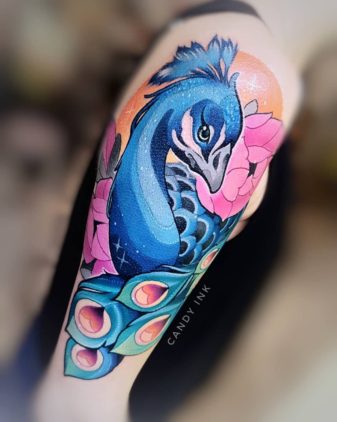 a neo traditional style tattoo of a peacock on the back of someone's arm. the peacock is facing to the left and is surrounded by pink flowers. the tattoo is done in a colorful palette with shades of blue, pink, orange, and green.