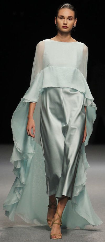 a full shot of a female fashion model on a runway wearing a light green, elegant gown with a flowing cape. the dress is made of a silky satinlike material, with a cropped top and wideleg pants. the model is wearing nudetoned high heels. her hair is slicked back, and she has a neutral makeup look. the background is black, emphasizing the pastelcolored attire. the overall ambiance of the photo suggests a highfashion event or runway show.