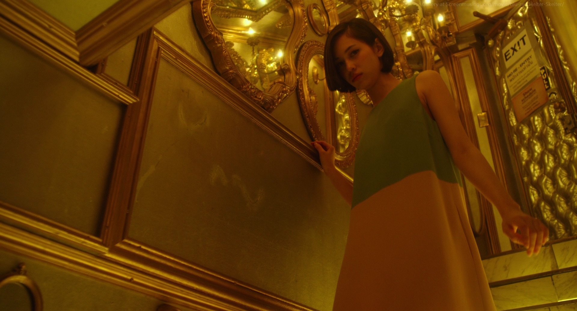 a woman stands in a vintage hallway with a dress that is green on top and light pink on the bottom. she is wearing a gold necklace and has her hair pulled back.  she is looking up and the hallway is ornately decorated with mirrors and gold trim.  an exit sign hangs on the door at the end of the hall.