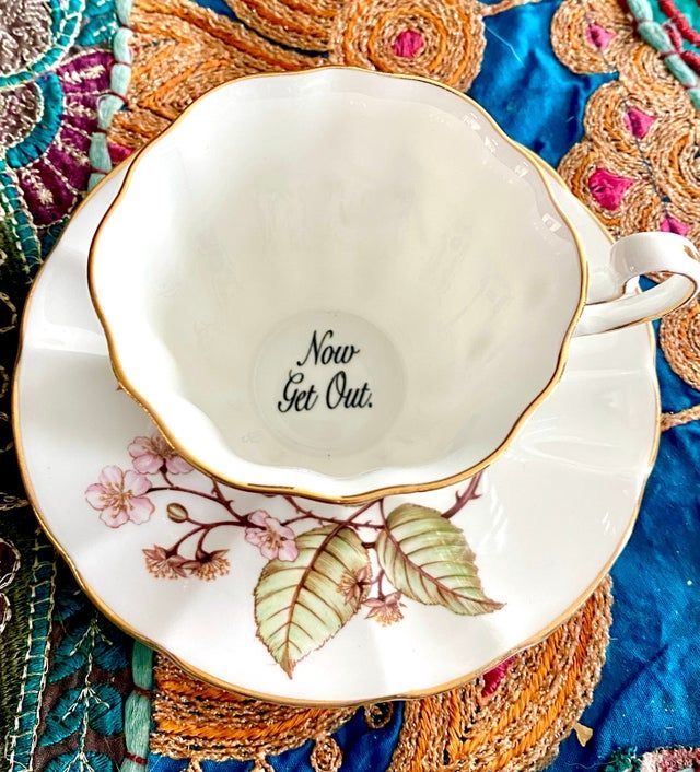 a white teacup with gold trim sits on a matching saucer. the saucer is decorated with a branch of cherry blossoms and leaves. inside the teacup, the message "now get out" is printed. the teacup and saucer sit on a colorful, decorative background.