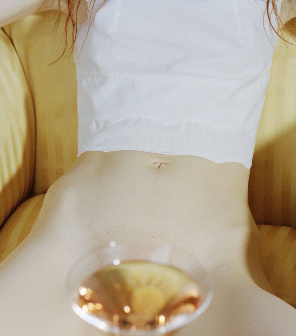 closeup view of a young woman's torso and abdomen. she is sitting on a yellow couch, wearing a white, loosefitting top that reveals her midriff and part of her lower abdomen.  a cocktail glass with a golden liquid is in the foreground, partially obscuring her lower body. the image has a soft focus and sensual quality. the overall feel is intimate and suggestive. the woman's skin is pale and her hair is partially visible.