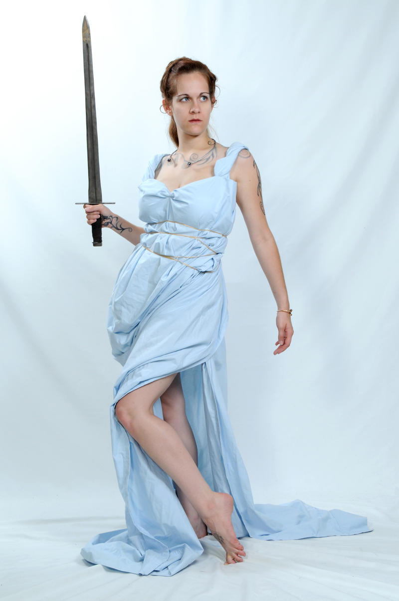 a woman stands on a white background in a studio setting, holding a sword. she is barefoot and wearing a light blue dress that is cinched at the waist with a thin rope. she has jewelry around her neck and a tattoo on her arm. she is posing in a warriorlike stance, looking off to the side with a determined expression.