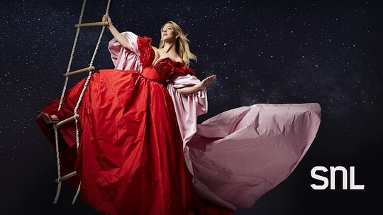 the image is a promotional still for saturday night live (snl), featuring a woman, likely the episode's host, in a dramatic pose. she is wearing a flowing, voluminous red gown with a lighter pink cape or overskirt that trails behind her.  she is perched on a rope ladder against a dark, starry backdrop. the overall aesthetic is elegant, slightly surreal and evokes a fairytale or fantasy theme. the snl logo is prominently displayed in the lower right corner. the pose and attire suggest a sense of glamour and perhaps a story or theme related to the episode.