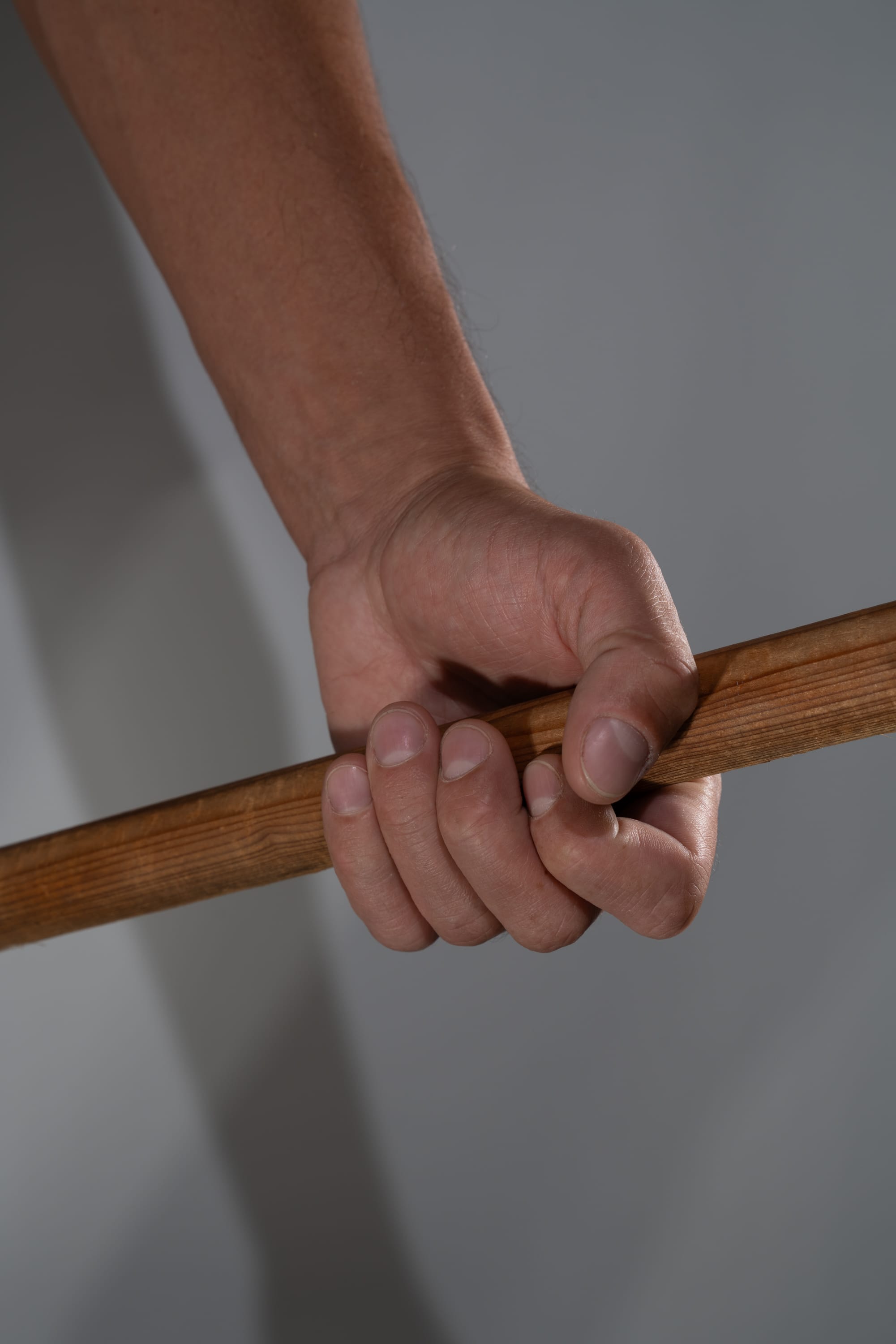 a closeup shot of a man's arm and hand firmly gripping a wooden stick or rod. the perspective is from below, emphasizing the strength and tension in the hand. the lighting is soft and even, highlighting the texture of the wood and skin.