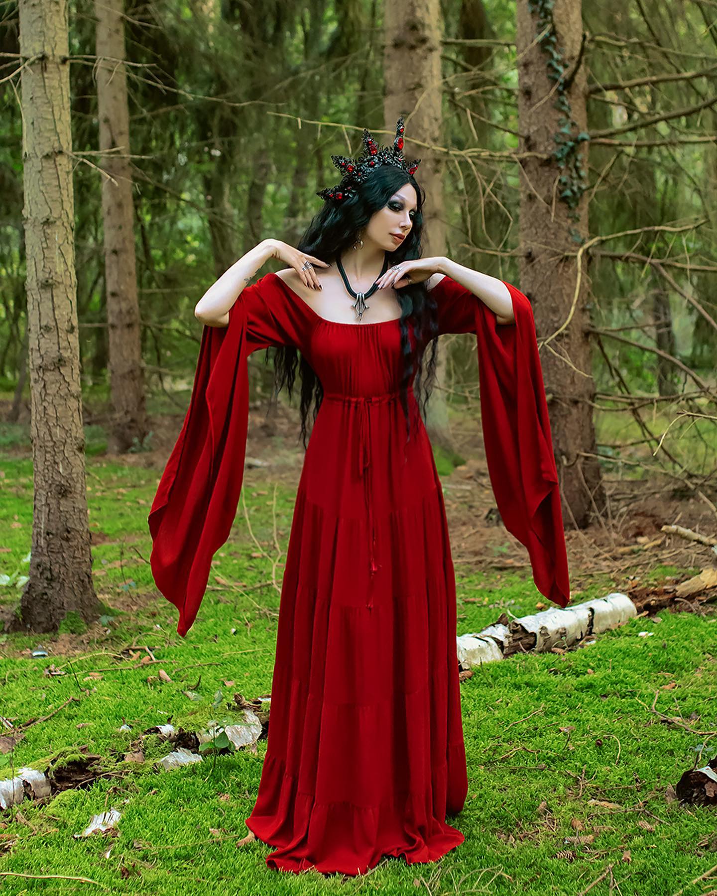 young woman with dark hair and a dark crown, wearing a long, flowing red dress with long sleeves, stands in a forest.  the setting is lush and green with moss covering the ground and tall trees surrounding her. the overall aesthetic is gothic, dark fantasy, and reminiscent of fairytale or medieval imagery. the woman's pose and expression are mysterious and elegant. the photo is high quality and welllit, capturing details in both the clothing and the natural environment.
