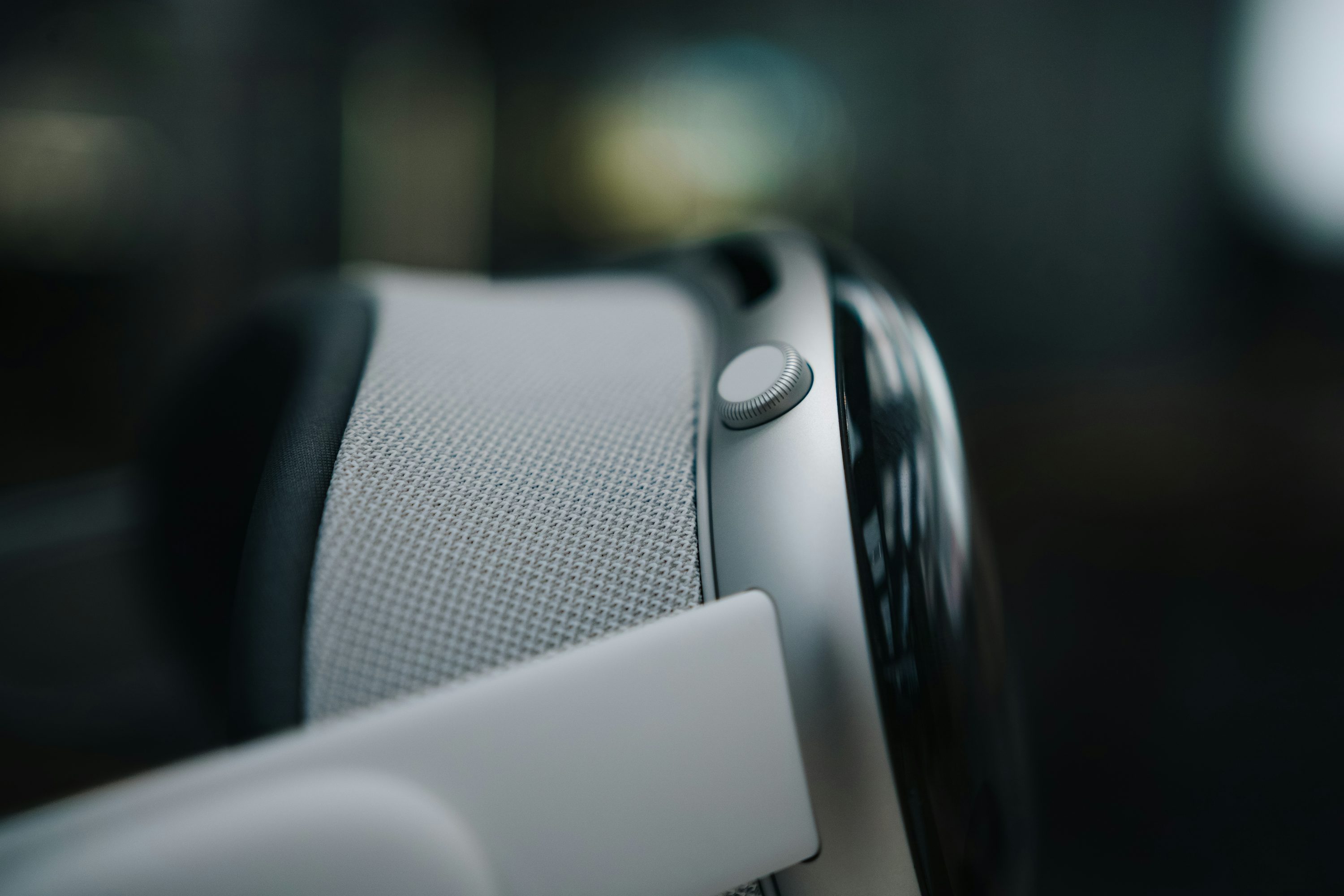 a closeup shot of the apple vision pro vr headset. the focus is on the side of the device, showcasing the digital crown, a grey fabric portion of the headband, the metallic frame, and some white components. the background is blurred.