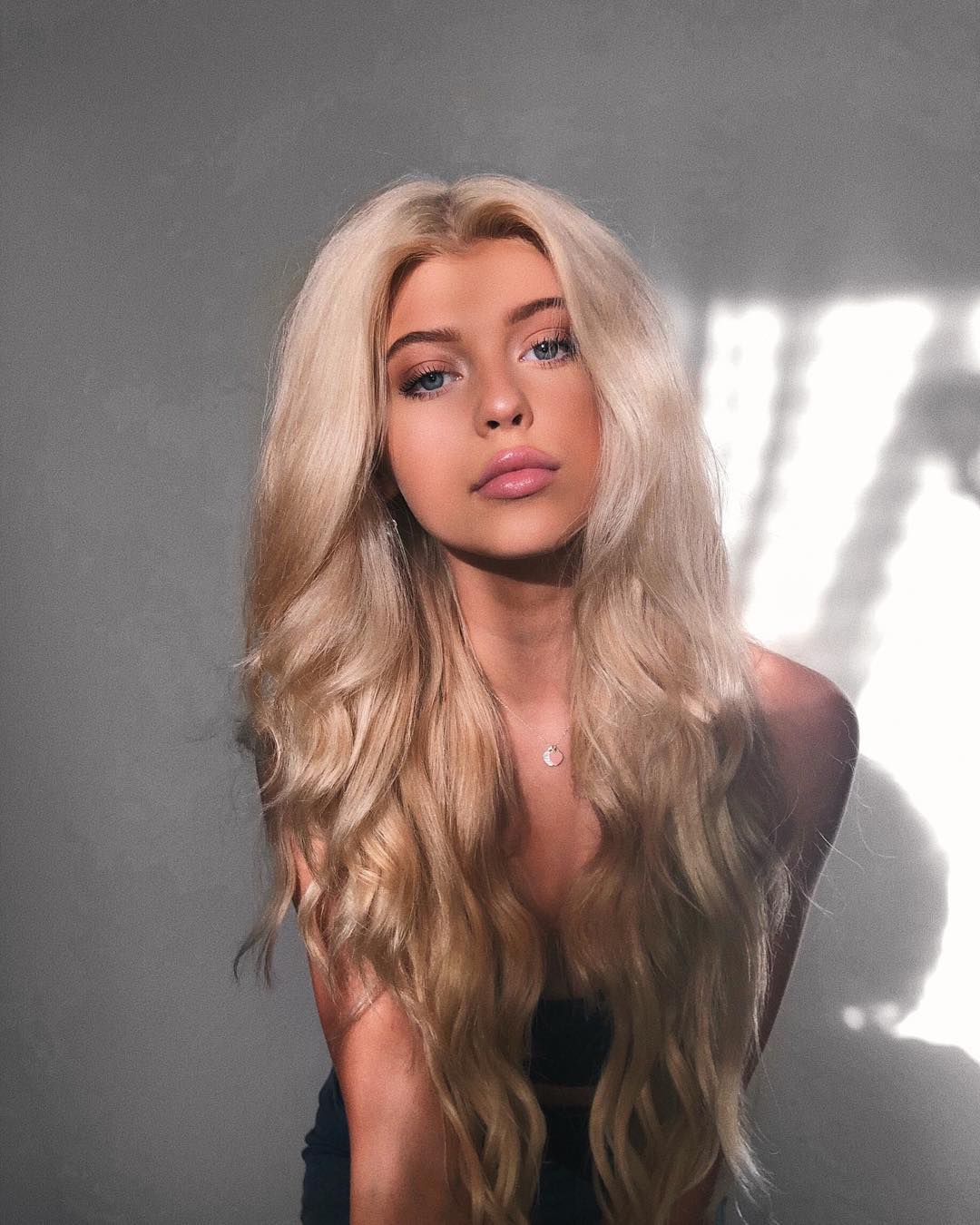 a portrait of a young woman with long blond hair and blue eyes. she is wearing a necklace. the background is a grey wall with a window shadow.