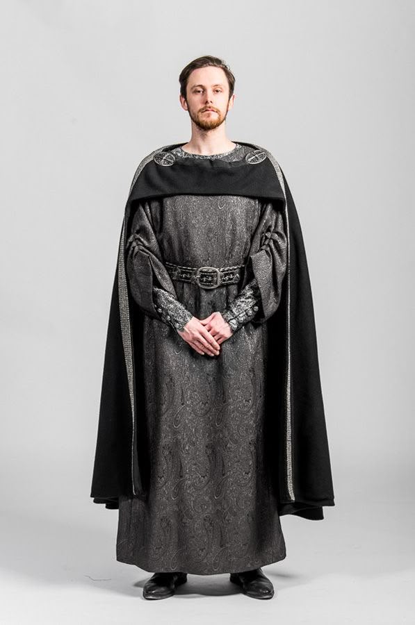 a man stands in a fulllength portrait, clad in a medievalstyle costume. he wears a long, intricately patterned grey robe cinched at the waist with a decorative belt. over the robe, he has a black cape, which drapes around his shoulders. he sports a neatly trimmed beard and looks directly at the camera. his footwear consists of shiny black shoes. the overall aesthetic suggests a historical or cosplay theme. the background is a plain, light grey color.