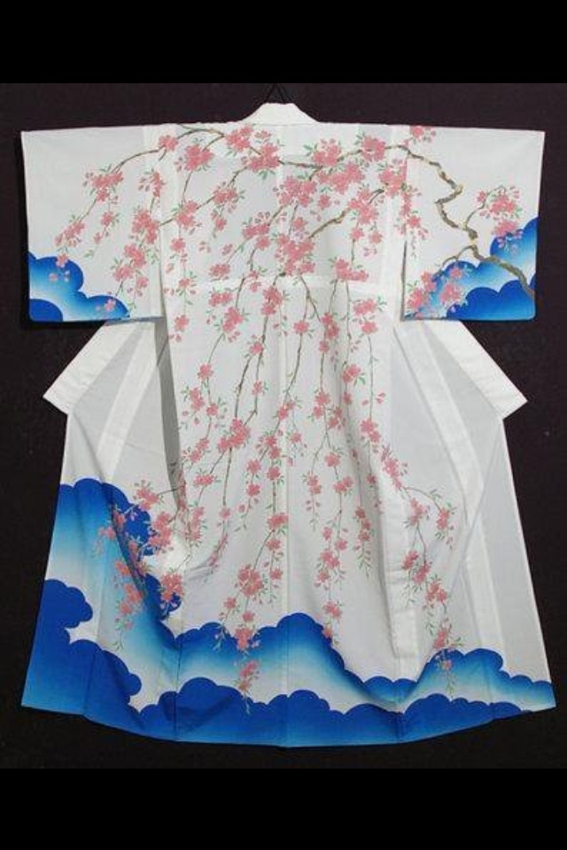 a full shot of a white kimono decorated with pink cherry blossoms and blue clouds, hanging in front of a dark background. the floral design drapes down the center and across the shoulders of the garment, while the blue clouds are located at the bottom and on the ends of the sleeves.