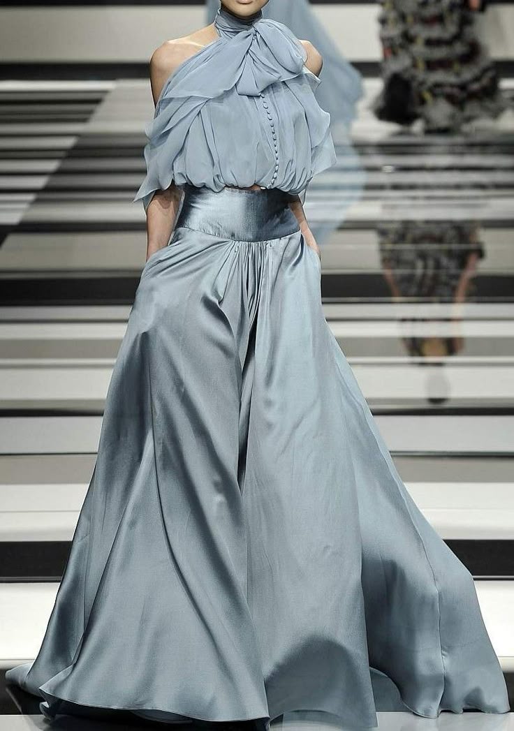 a woman is standing on a runway wearing a blue silk gown. the gown features wide leg pants and a halter top with an offtheshoulder design. the runway is black and white.