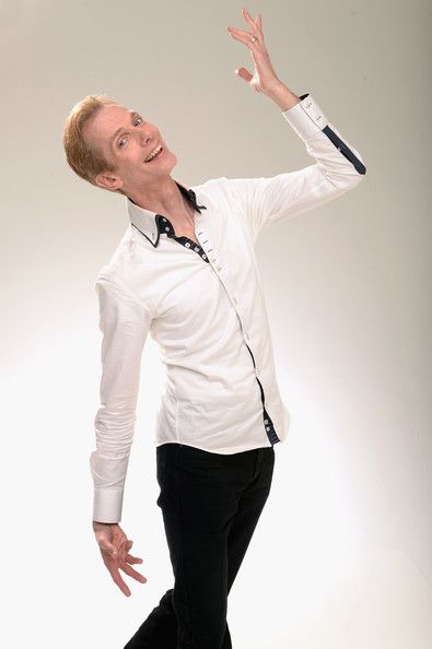 a man is standing in front of a white background. he is wearing a white shirt and black pants. he is smiling and looking up. he is raising his arms. the man is dressed stylishly and looks confident and happy.