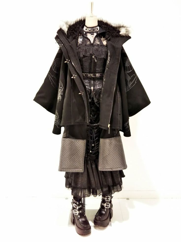 a fashion model is wearing a black gothic outfit. she's wearing a black jacket with a faux fur hood. the jacket has decorative embroidery on the arms. she's also wearing a black lace top, a skirt with layered fabric, and  black boots with buckles.