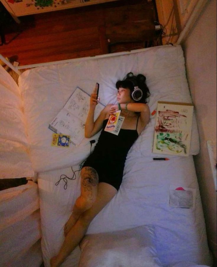 a woman is lying on a white bed in a bedroom. she's wearing black tank top and headphones and holding a cell phone. she has a tattoo on her leg. there is a drawing on the bed and a book beside her. the woman looks relaxed and comfortable.