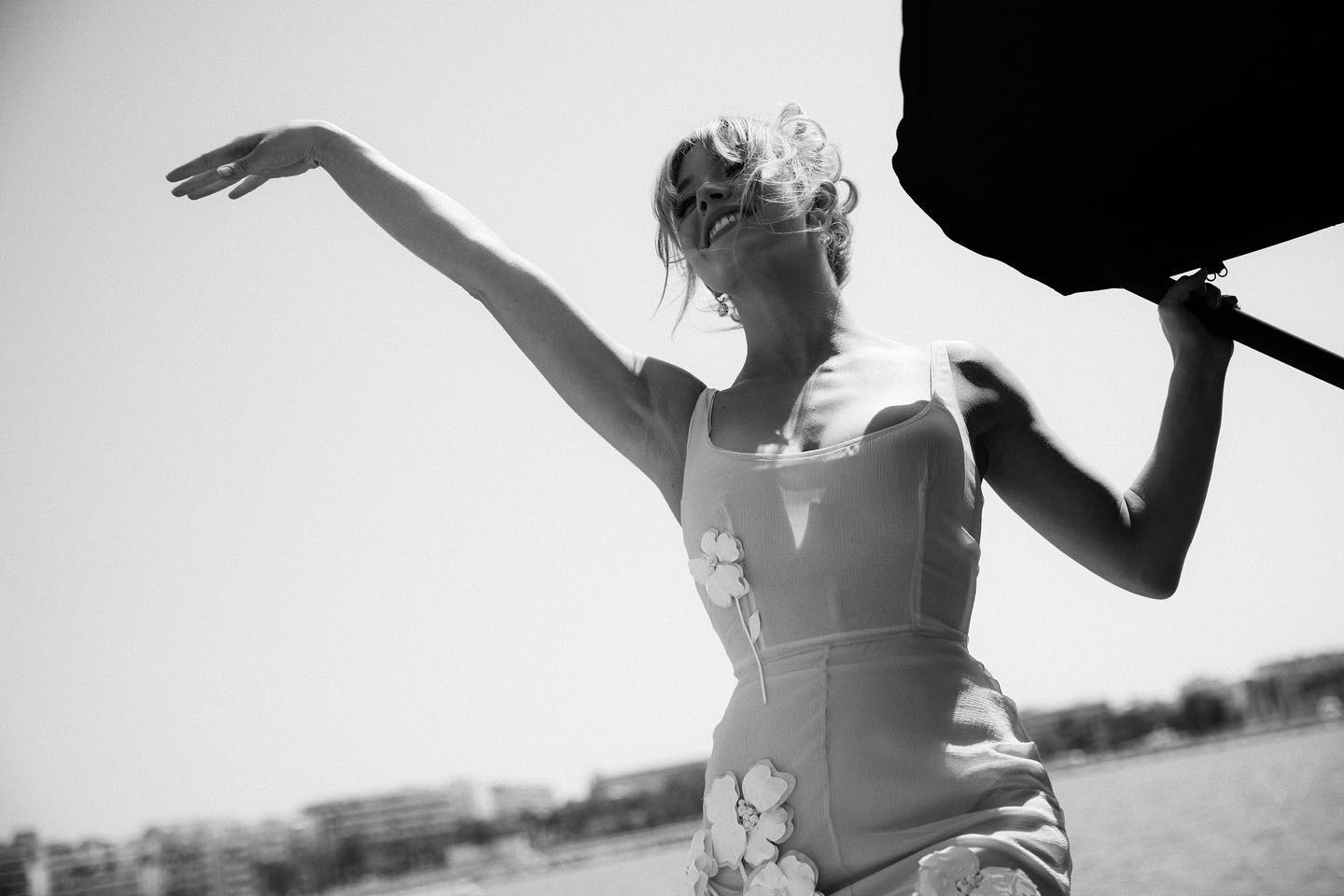 a black and white, lowangle shot captures a woman in a lightcolored, floral summer dress. her arm is elegantly raised, and she is holding a black reflector.  the background is slightly out of focus, suggesting a beach or seaside setting with buildings in the distance. the overall mood is bright, sunny, and joyful, creating a candid yet stylish fashion portrait.