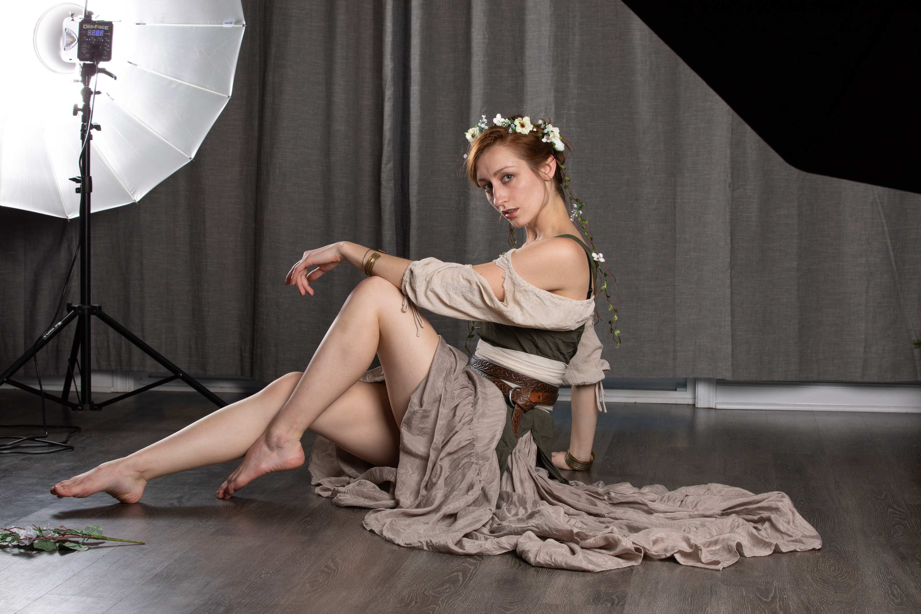 a woman in a flowing, light brown dress poses elegantly on a dark wooden floor in a photography studio. she wears a flower crown and simple jewelry, her pose highlighting the drape of her dress. a professional softbox light illuminates her from the left, casting a soft light on her and creating a contrast between light and shadow. the background is a simple dark grey curtain, keeping the focus on the model and her attire.