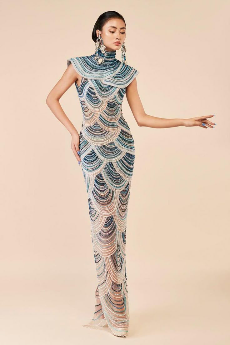 a slender asian woman models a striking couture gown with an elaborate beaded design. she poses elegantly against a neutral backdrop, her arm extended gracefully. the gown, reminiscent of mermaid scales, features a high collar and a formfitting silhouette. statement earrings add to the overall glamorous look.