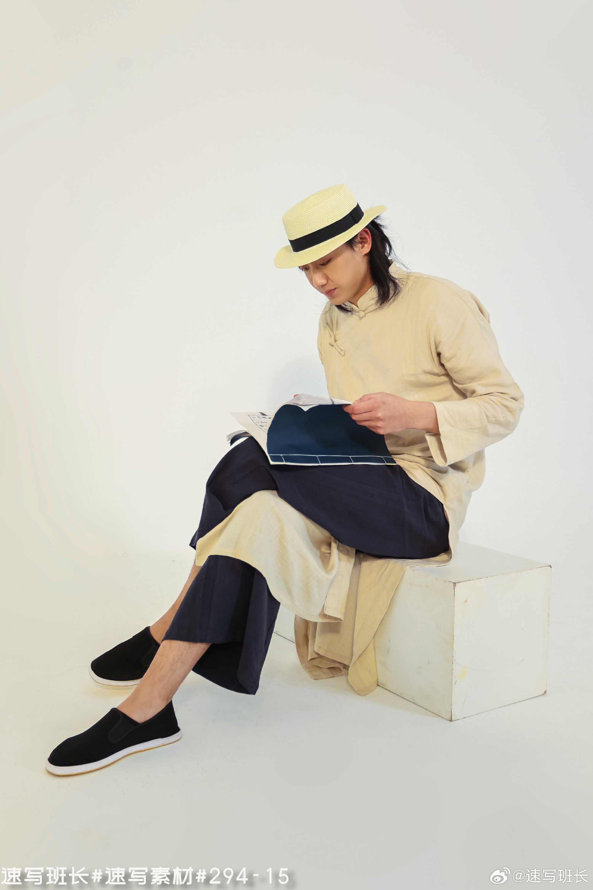 a young man wearing a beige hanfu with black pants and black cloth shoes sits on a white cube, reading a magazine. he has long black hair tied back and wears a straw hat. the background is plain white, and the image appears to be from a photoshoot.