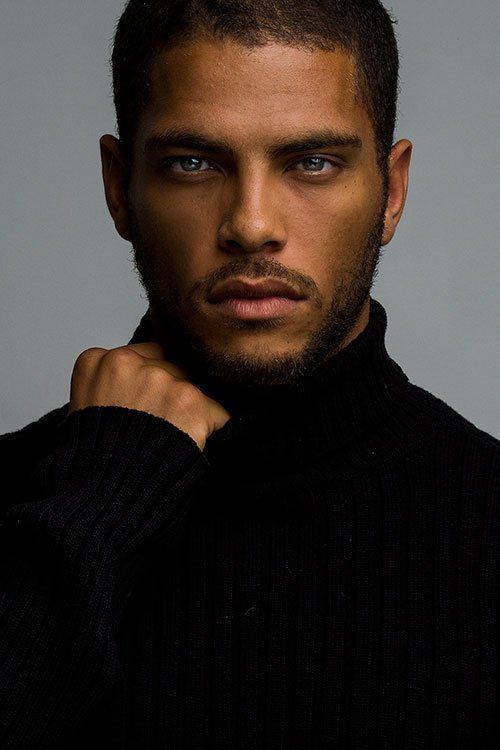 a closeup studio portrait of a handsome darkskinned man with short hair and a neatly trimmed beard, wearing a black turtleneck sweater. he has striking blue eyes and a serious expression. his hand is slightly raised, touching his neck. the background is a plain gray.