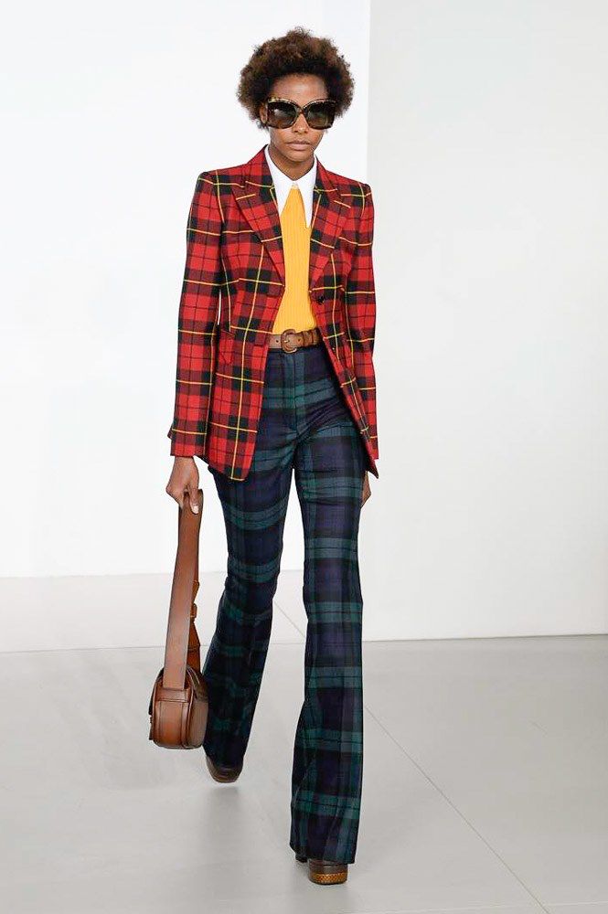 a woman walks down a runway wearing a red and black plaid blazer over a yellow top and blue and green plaid pants. she is wearing sunglasses and carrying a brown bag. she is also wearing brown shoes.