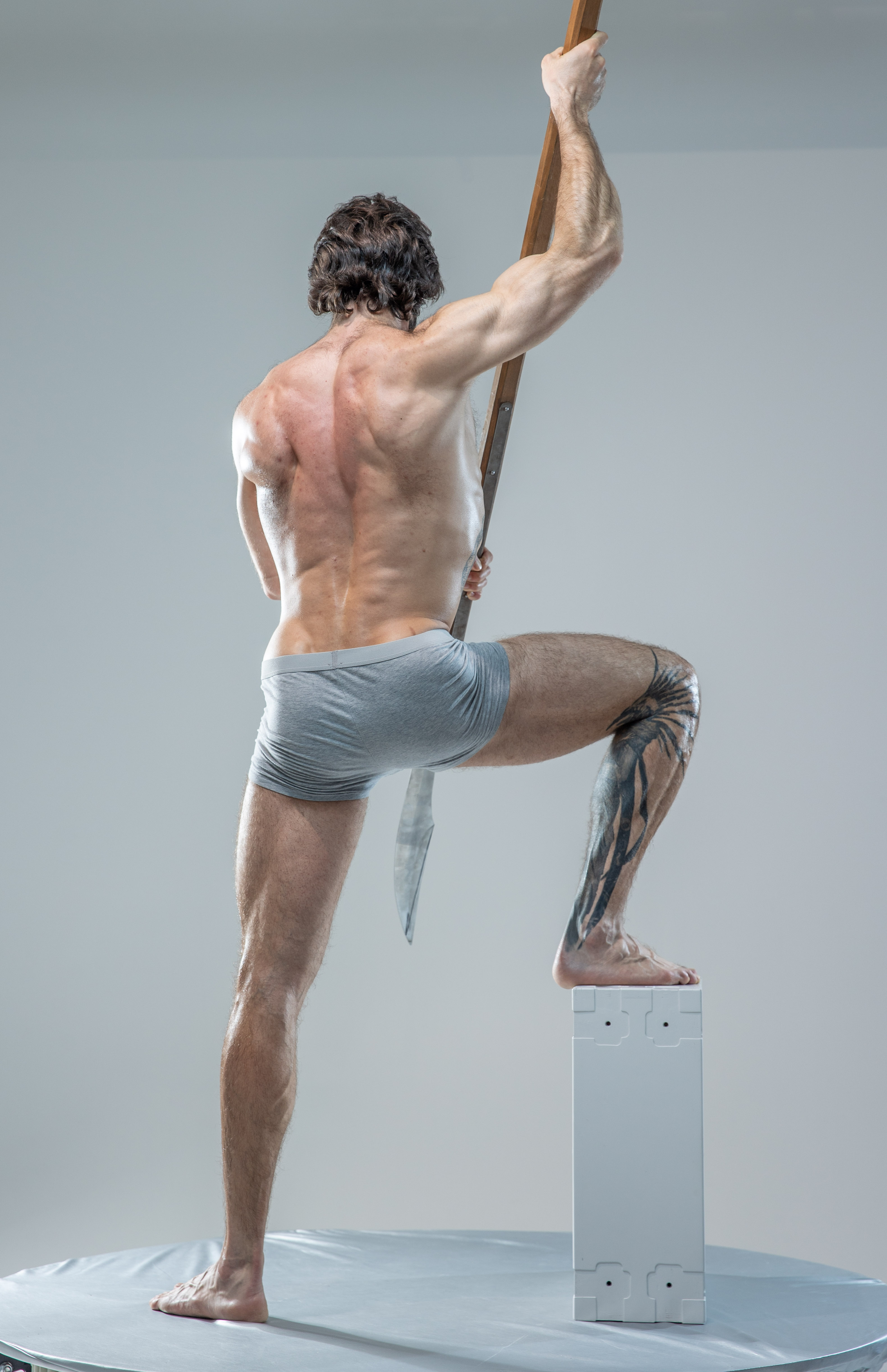 a male model with long hair is standing on a small white platform, facing away from the camera. his left leg is bent, resting on a white cube. he holds a wooden staff diagonally in his right hand, extending it behind him. the background is a solid gray, creating a studiolike setting. the lighting is soft, highlighting the contours of his muscles.