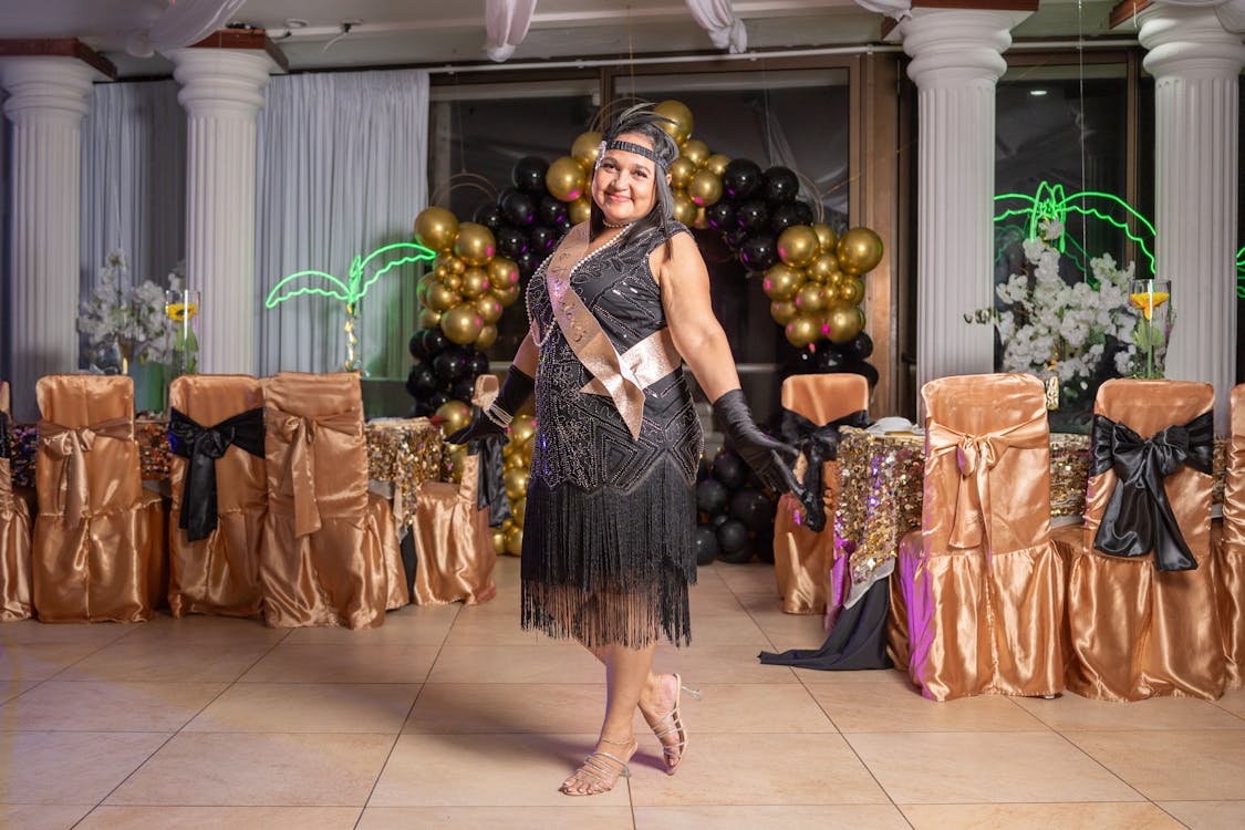 a woman dressed in a black flapper dress, headband, and gloves stands smiling at her 30th birthday party. she wears a birthday sash that says "30". the party is decorated with gold and black balloons, columns, and tables with goldcovered chairs. a palm tree light provides a tropical touch.