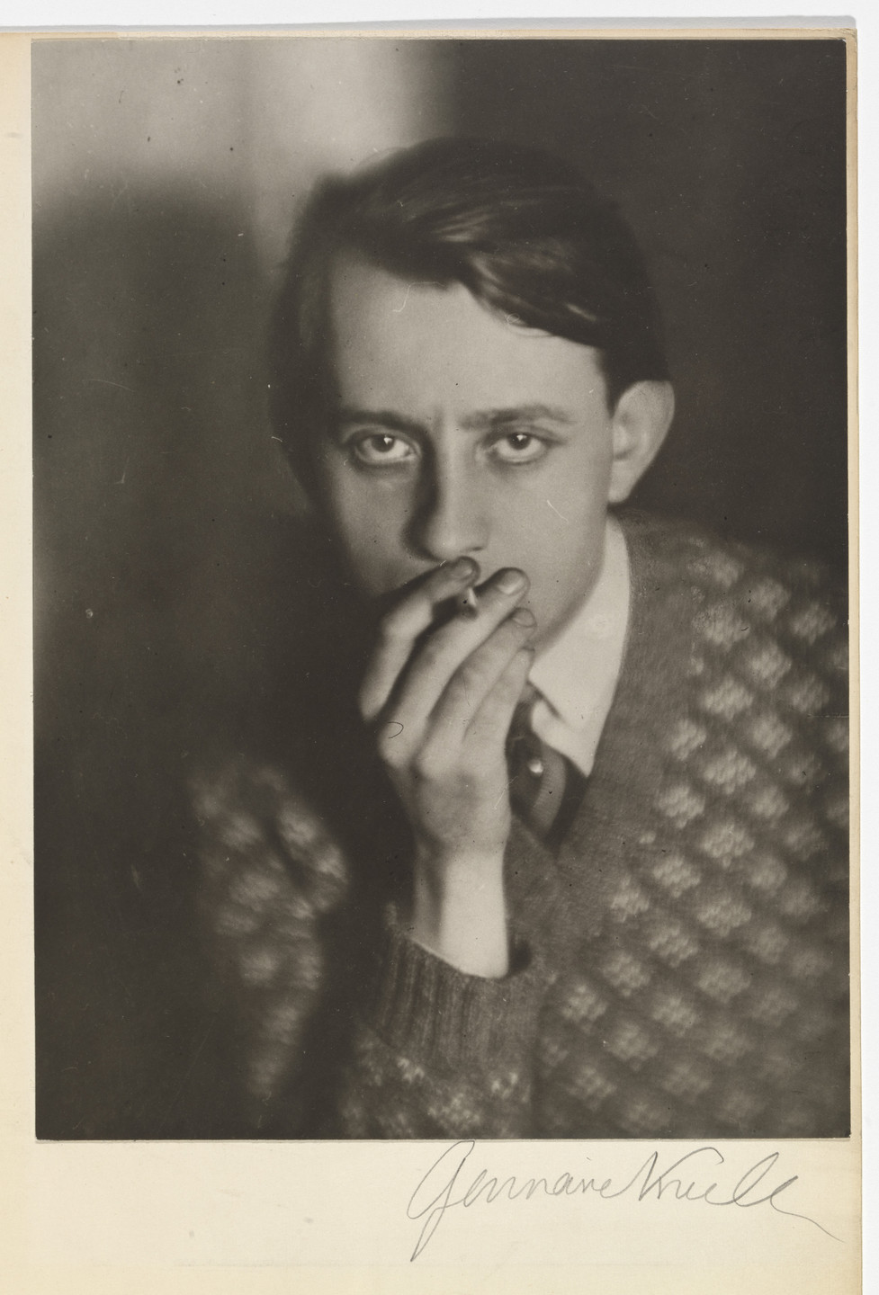 a black and white portrait photograph of a young man, possibly in his twenties, who appears to be smoking a cigarette. he is wearing a patterned sweater and a collared shirt, and his expression is serious and pensive. the photograph is a closeup shot that focuses on his face and upper body. the photograph is signed "germaine krull" in the bottom right corner. the style of the photograph is consistent with the new objectivity movement in photography. the image has a slightly dark and moody atmosphere, but also has a certain amount of sharpness and clarity which is typical of 20th century photography.