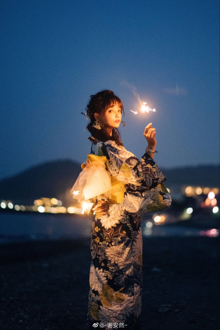 a young asian woman in a beautiful yukata stands on a beach at night, holding a lit sparkler. the background is blurred, showing a cityscape of lights across the water. the overall atmosphere is romantic and serene.  the woman's expression is calm and content. the image is welllit, capturing the sparkle of the fireworks and the soft light on her clothing.