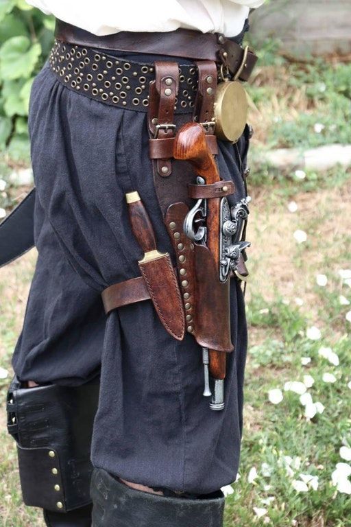loseup view of a person wearing a pirate costume. the focus is on a detailed leather holster and belt system attached to their hip. the holster contains a flintlock pistol and a knife. the belt is adorned with metal studs and features a small, round, brasscolored container. the person is wearing darkcolored breeches and a white shirt.  the background is blurred but shows green grass. the overall impression is one of historical or fantasy reenactment.