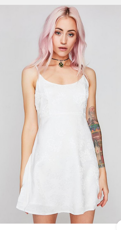 a young woman with pink hair and tattoos is modeling a white mini dress. the dress has thin straps and a floral pattern. the background is a light gray.