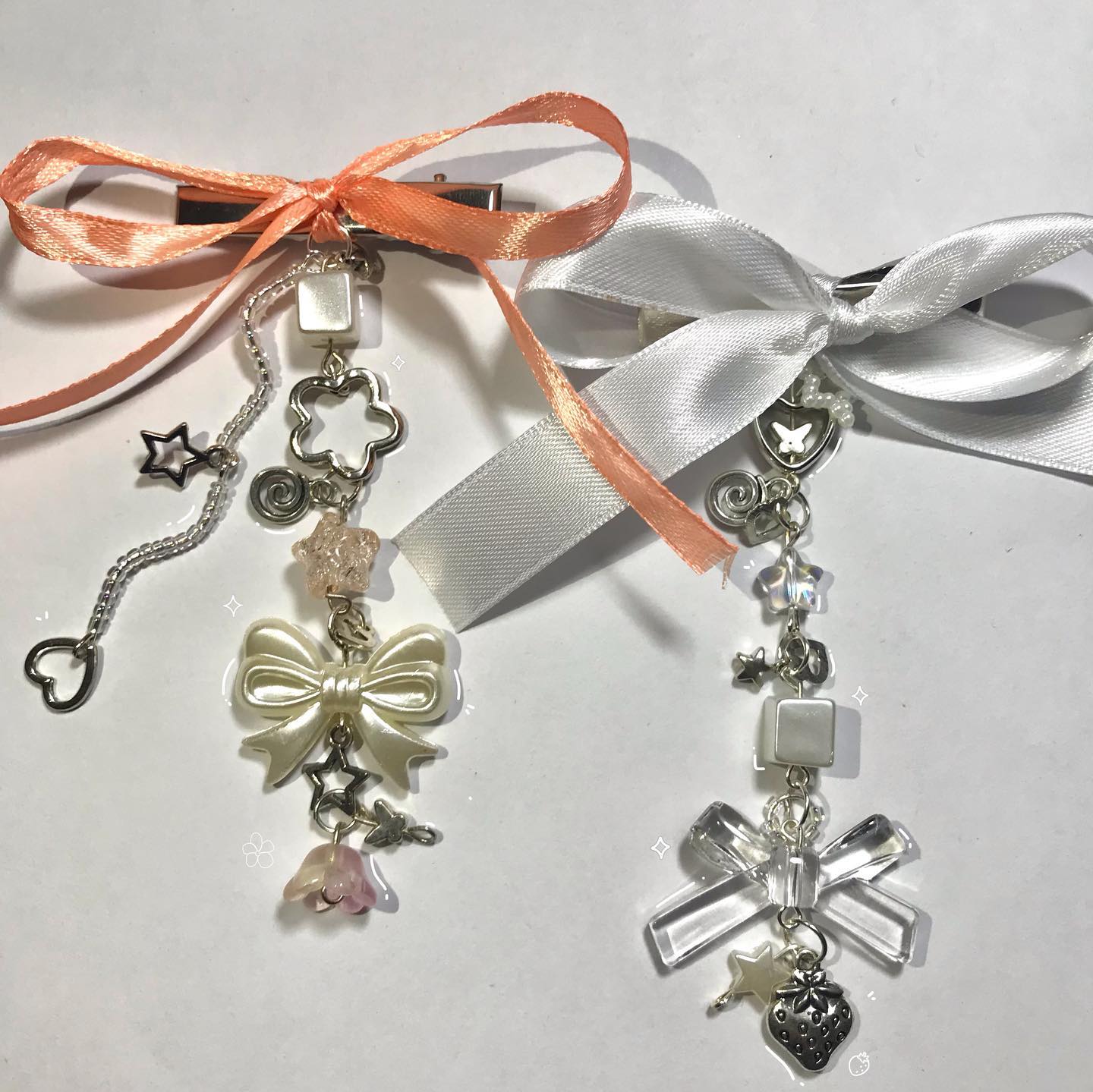 two hair clips with bows and various charms are displayed on a white surface. the clip on the left features an orange ribbon bow and charms like a heart, star, flower, and a bow charm. the clip on the right has a white ribbon bow and charms including a strawberry, star, butterfly, and bow charm.