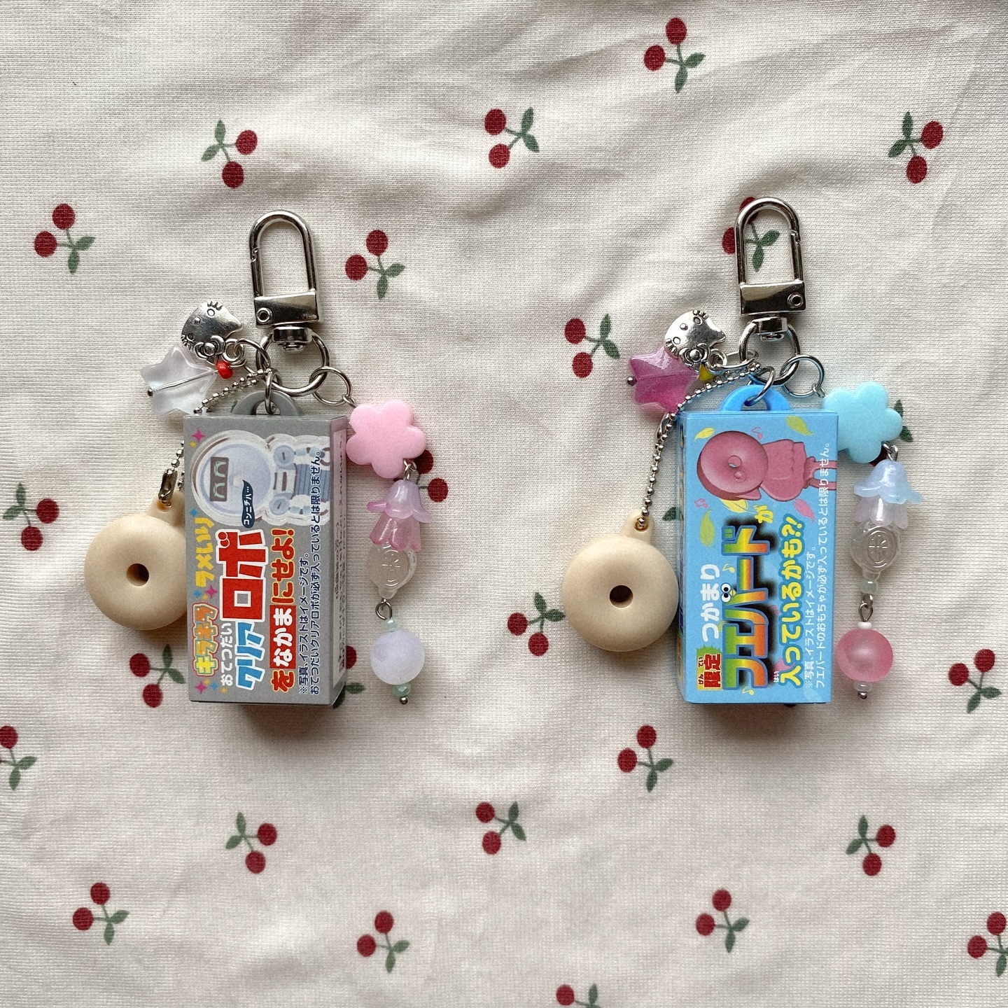 two keychains lie on a cherrypatterned surface. each keychain has a silver clasp attached to a charm resembling a small box with japanese text. one box is gray, and the other is blue. additional charms, including a donut, a flower, and beads, dangle from each keychain. the background features a repeating pattern of small red cherries with green stems on a creamcolored fabric.