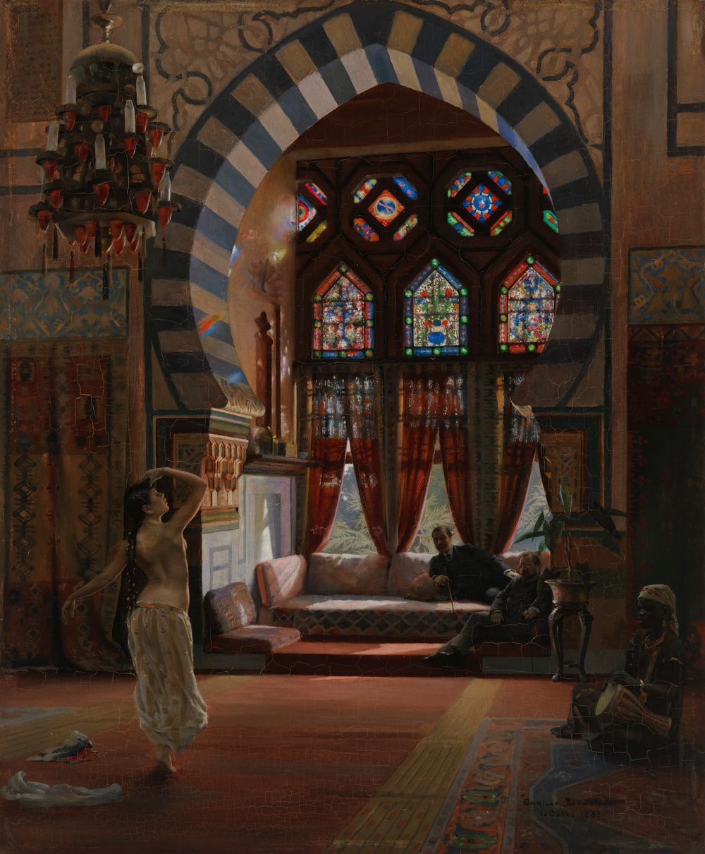 an orientalist painting depicts a room with a large archway leading to a window with stained glass. inside the room, a woman with bare shoulders and a veil dances. two men are seated on a sofa, while another man with a turban and drum sits on a rug in the corner. a chandelier hangs from the ceiling, and the room is decorated with an ottoman rug and cushions.