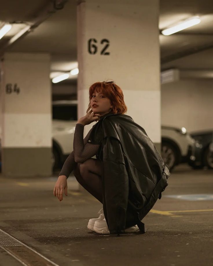 a young woman with red hair is squatting in a parking garage, wearing a leather jacket and sneakers. she is looking to the side, with a cool and stylish expression.