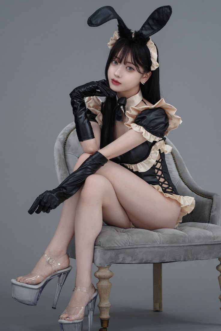 a woman dressed in a bunny girl cosplay outfit sits in a gray chair. she has black hair, bunny ears, and is wearing black leather gloves and a black dress with lace details. she is also wearing clear platform heels. the background is a plain gray color. she has her right hand by her face as if posing.