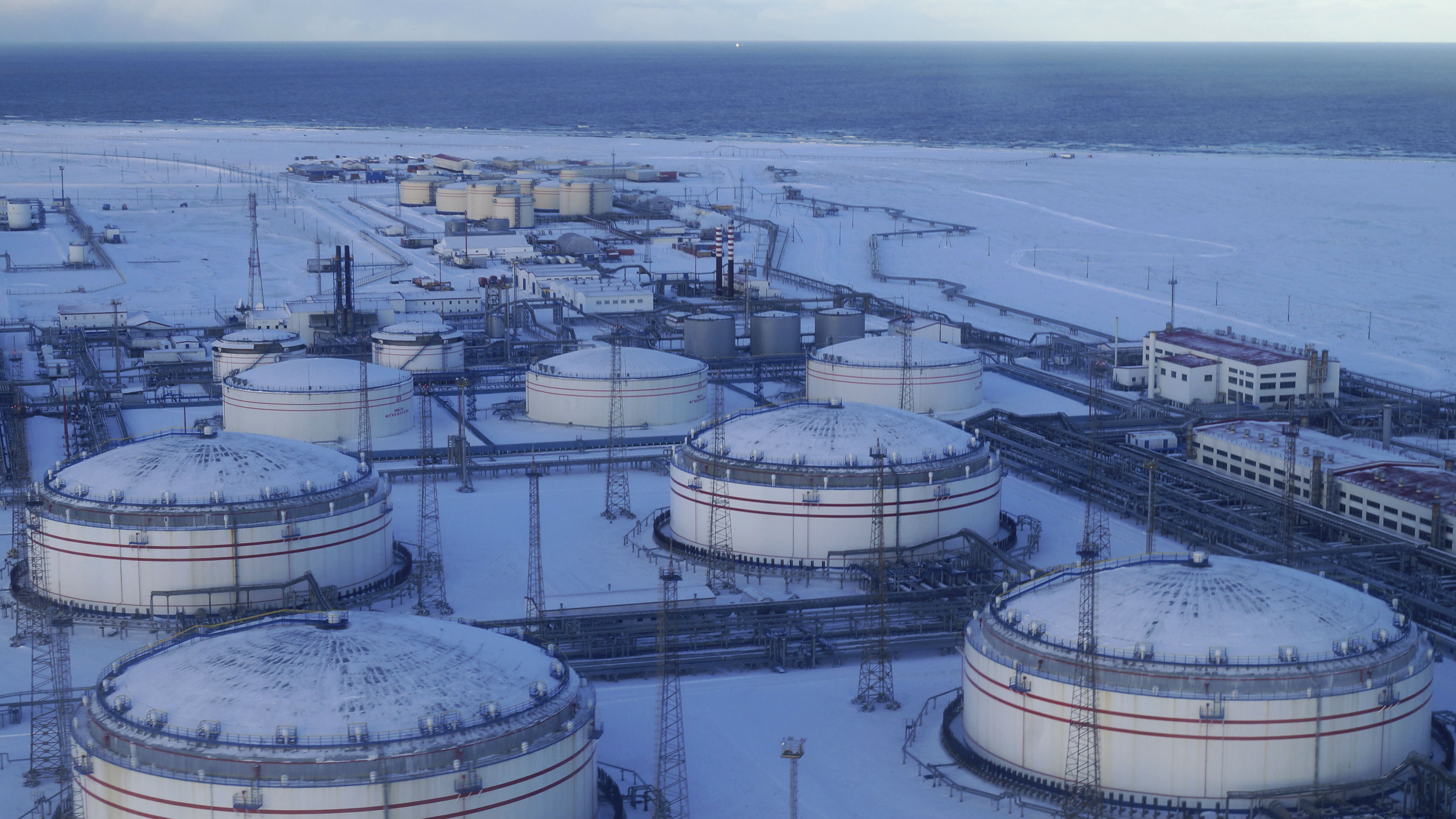 a wide aerial shot of an oil refinery in the arctic. the refinery is covered in snow, and there are numerous large oil tanks, pipelines, and other industrial structures. in the background, you can see the frozen ocean. the photo highlights the harsh environment and the vast scale of oil production.