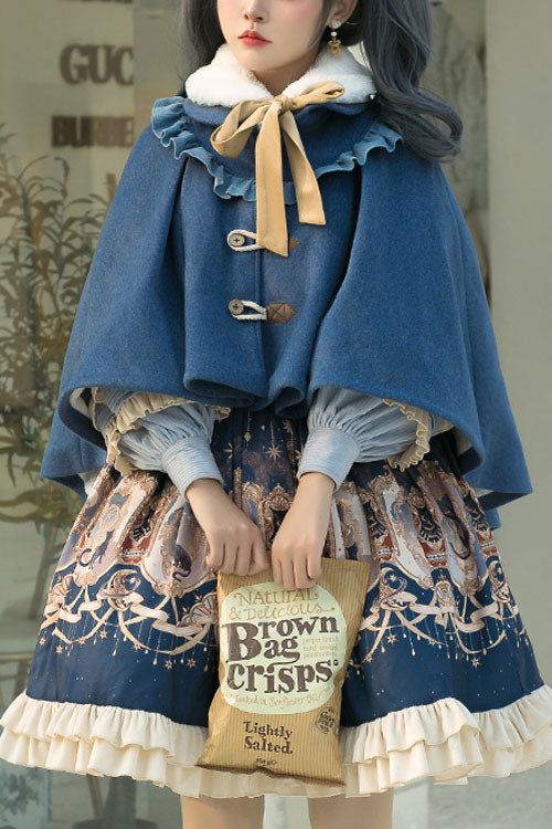 a woman is wearing a blue wool cape with a faux fur collar and a blue and cream lolita dress with a celestial print. she is holding a bag of crisps in her right hand. the image is well lit and the woman is the focus of the image.