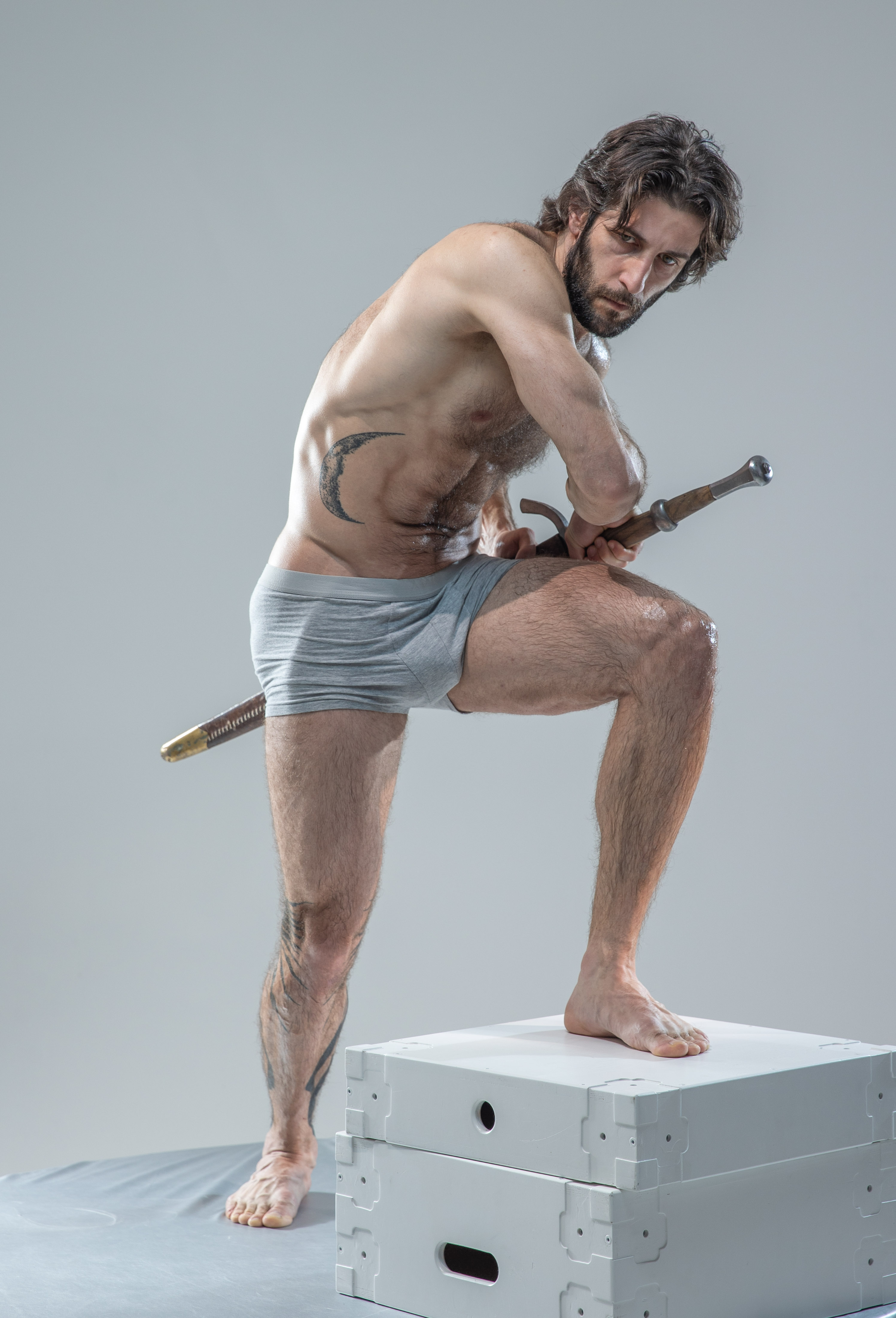 a muscular man with a beard and long hair is standing on a white box in a studio. he is holding a sword and is wearing gray underwear. he has a tattoo on his left hip. the background is gray.