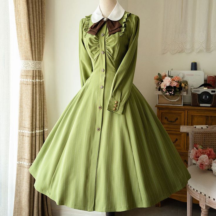 a green vintagestyle dress is displayed on a mannequin. the dress features long sleeves, a collar, a row of buttons down the front, and a full skirt. a brown bow tie completes the look. the background provides a glimpse into a welllit room with floral arrangements and a vintage sewing machine.
