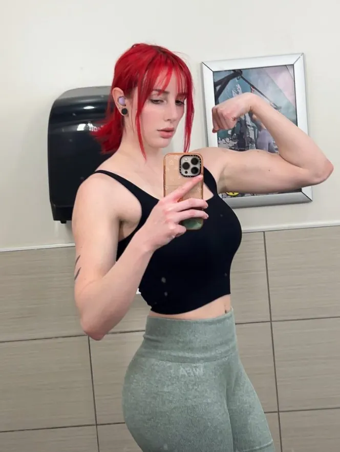 a young woman with vibrant red hair and bangs is taking a selfie in a gym bathroom mirror. she's flexing her biceps and showing off her athletic physique. she is wearing a black sports bra,  sage green leggings and appears to be wearing earbuds. the mirror reflects the bathroom tiles and a framed picture on the wall behind her.  the overall impression is one of fitness, strength, and selfconfidence.
