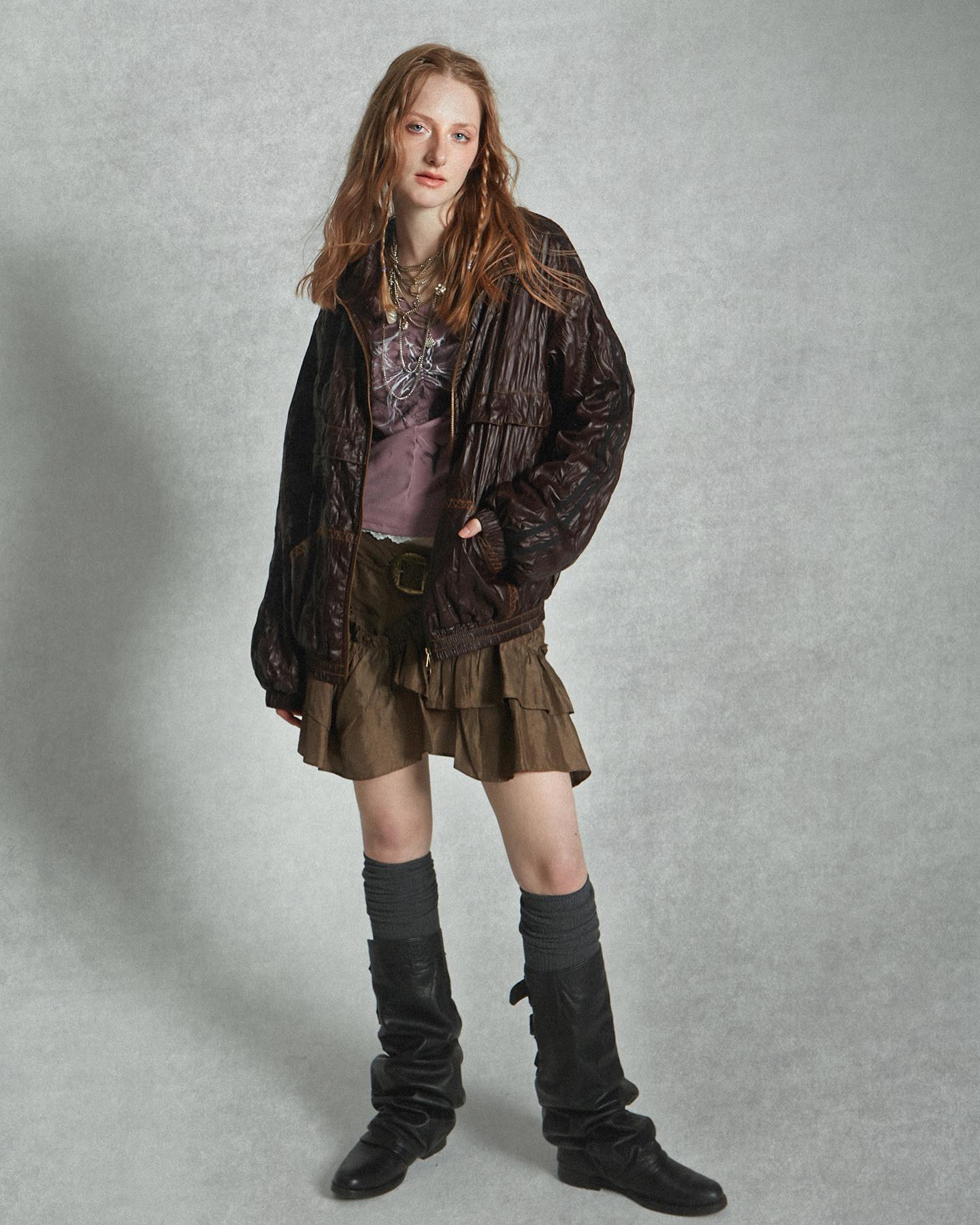a fulllength studio shot of a young woman, seemingly a fashion model, posing against a neutral grey backdrop. she's dressed in a layered outfit that evokes a grunge or bohemian aesthetic: a dark brown, quilted jacket with a relaxed fit over a mauvetoned printed top; a layered, ruffled brown miniskirt; dark grey kneehigh socks, and slouchy black leather boots.  her hair is long and reddishbrown, parted down the middle, with a slightly undone look.  she wears several necklaces.  the overall impression is one of casual, slightly edgy style.