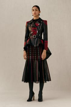 a woman wearing a black leather jacket with red plaid accents, a pleated black skirt with red stripes, and black high heels. she is posing against a creamcolored background.