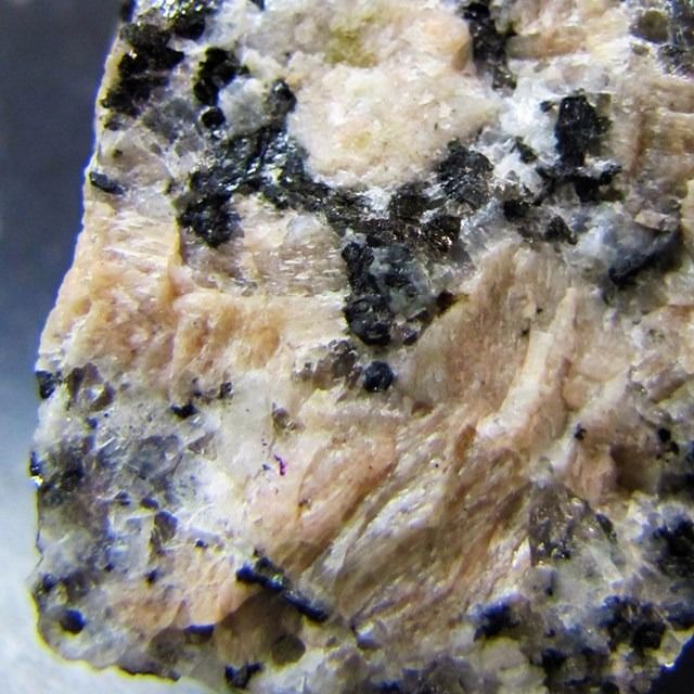 a closeup image of a granite rock, displaying its unique texture and crystalline structure.  the granite exhibits a heterogeneous mixture of crystals in varying sizes, shapes, and colors. some crystals are larger and more defined, while others are smaller and appear as a network of lines. the overall composition of the granite is lightcolored, with a slight pink hue, indicative of the presence of feldspar. black, specks of biotite can be seen throughout the granite, adding a dark contrast. the surface of the rock is rough and uneven, showcasing the natural formation of the rock. this image provides a visual representation of the intricate details found in a granite rock.