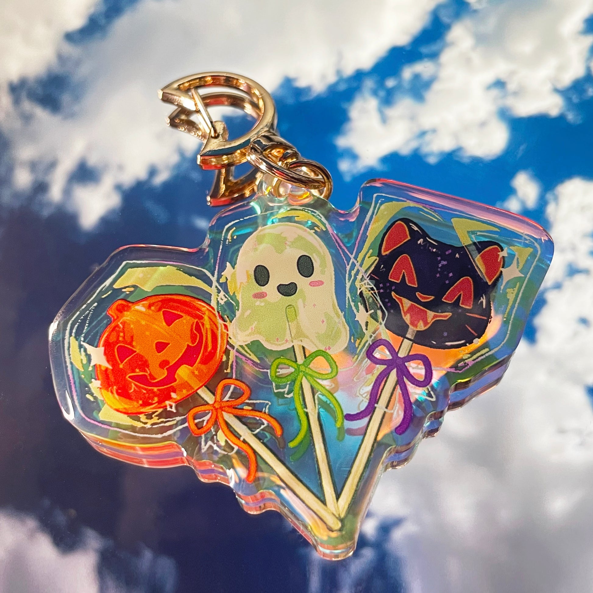 closeup view of a halloweenthemed keychain. the keychain is made of iridescent acrylic and features three lollipops: a ghost, a pumpkin, and a black cat. each lollipop has a unique design and is attached to a stick.  the keychain is shaped like a slightly irregular blob, and the background is a bright blue sky with fluffy white clouds.  a gold colored keyring is attached at the top. the overall style is cute and whimsical.