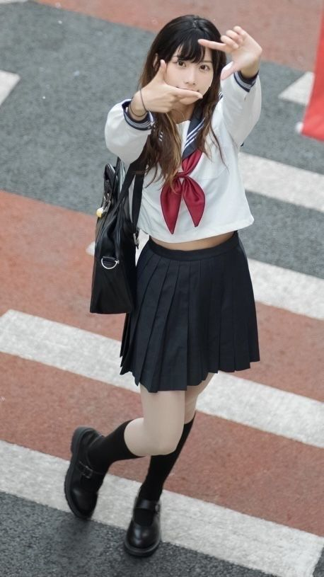 a young woman wearing a sailorstyle school uniform stands on a crosswalk. she is posing for the camera, making a framing gesture with her hands. she is carrying a black purse and wearing black socks and shoes. the background suggests a busy street scene.