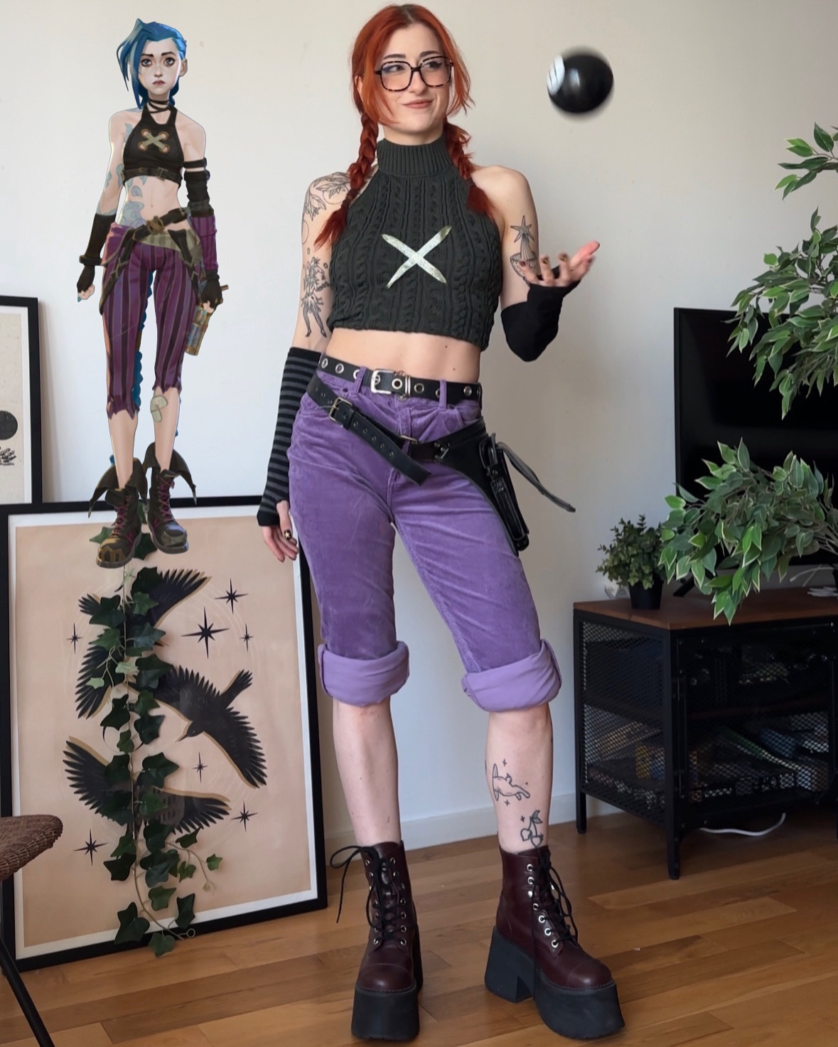 the image shows a young woman cosplaying as jinx from the video game league of legends.  she's dressed in a costume that captures jinx's style: a purple cropped corduroy pants, a dark green cropped cableknit halter top, and dark brown platform boots.  her accessories include black wristbands and a utility belt.  she has red hair styled in braids and is wearing glasses. she has visible tattoos on her arms and legs. a framed piece of artwork depicting birds and vines is visible on the wall behind her. she is standing in a home setting, seemingly juggling a small dark ball.  a depiction of jinx from the game is visible in a picture frame in the background for comparison.